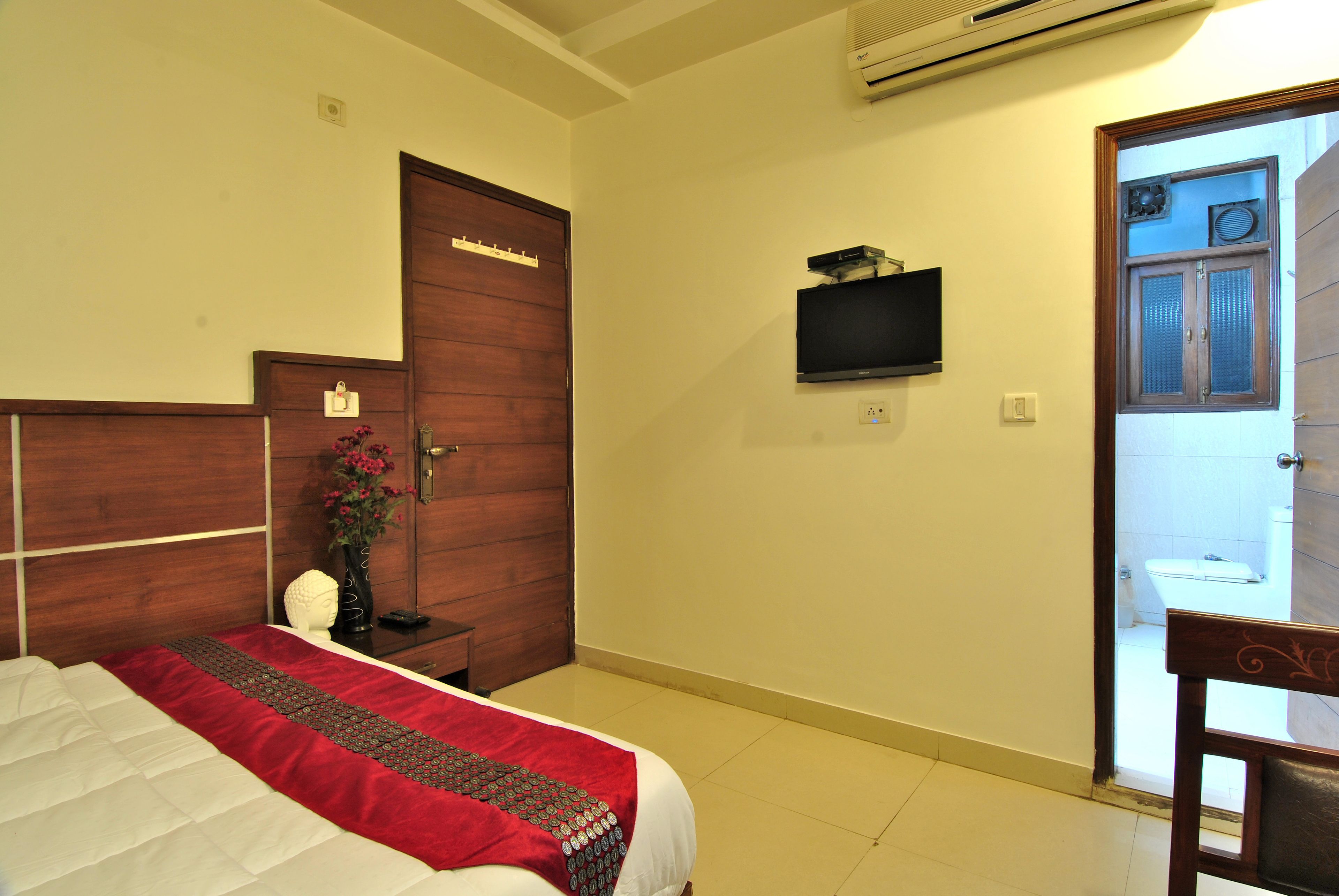 Habitación de hotel con una cama, una puerta de madera, un televisor montado en la pared y un baño visible a través de una puerta abierta.