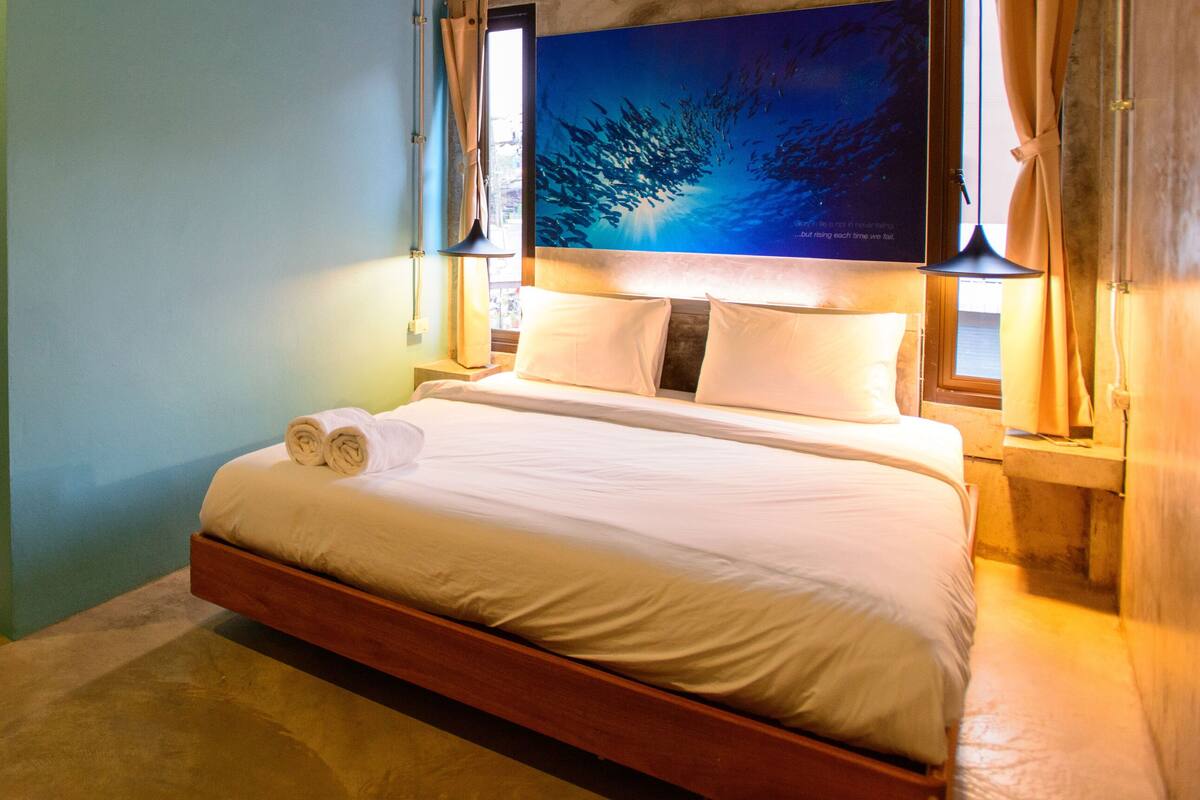 A hotel room with a bed, a painting, a window with curtains, and a bathroom visible through an open door.