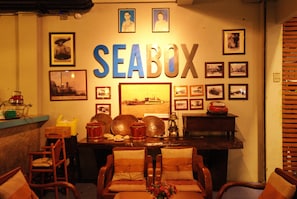 Una habitación con un letrero grande en la pared que dice "SEABOX", rodeada de fotografías enmarcadas y diversas decoraciones con temática marítima. Hay dos sillas de madera y una mesita con un jarrón de flores.