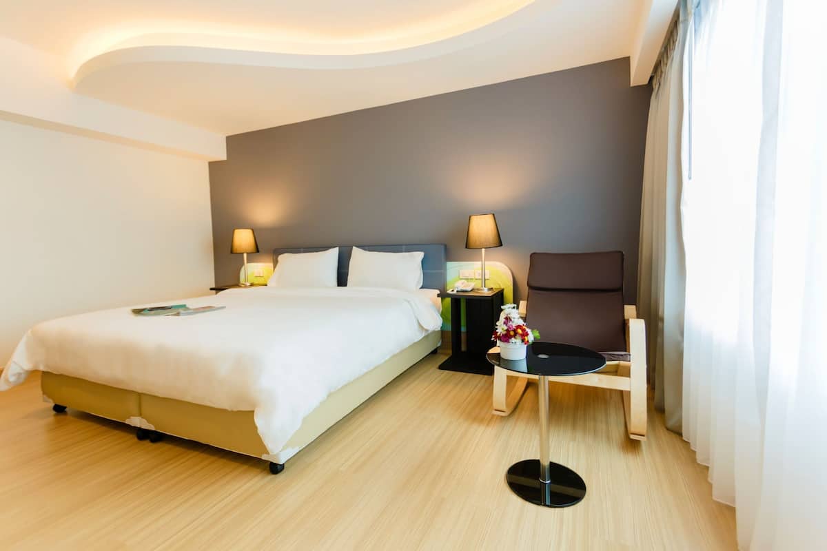 Quarto de hotel moderno com uma cama grande, duas luminárias de cabeceira, uma mesinha com um vaso de flores e uma cadeira.