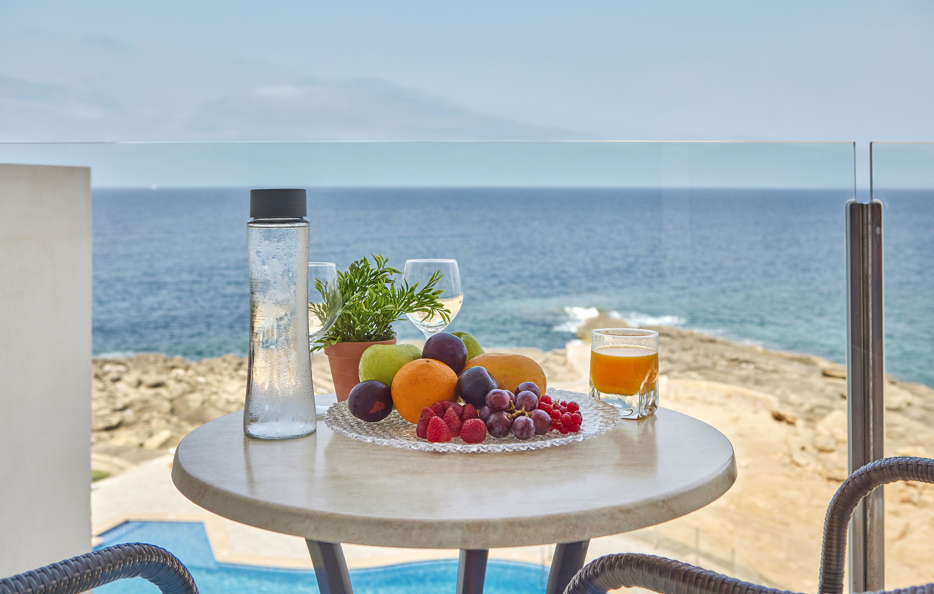 Un tavolo con frutta, un bicchiere di succo e una bottiglia d'acqua su un balcone vista mare.