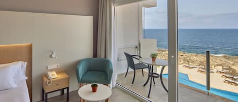 A hotel room with a bed, a desk with a phone, a chair, a small table with a plant, and a balcony with a pool and beach view.