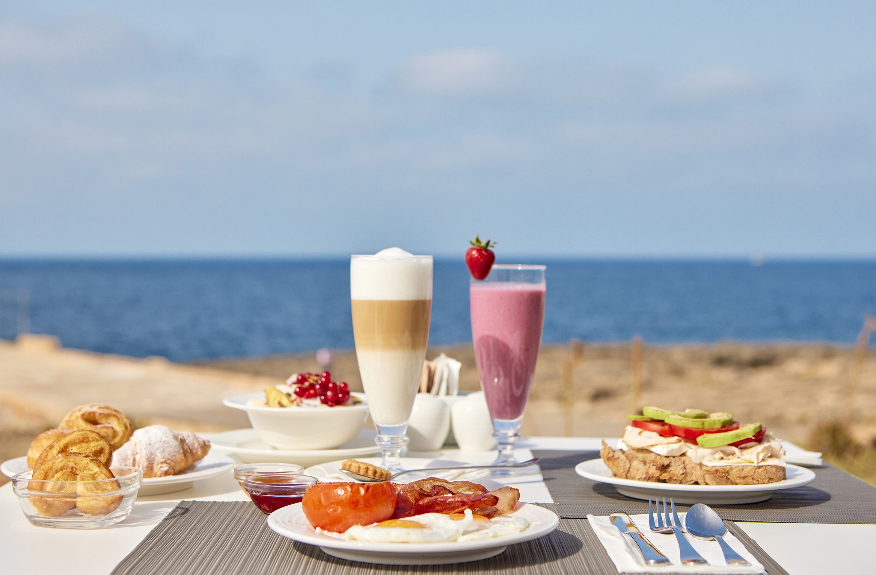 Una mesa dispuesta para el desayuno, con bollos, una bebida, y un plato de huevos y tocino, con vista al mar.
