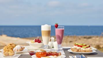 Una mesa dispuesta para el desayuno, con bollos, una bebida, y un plato de huevos y tocino, con vista al mar.