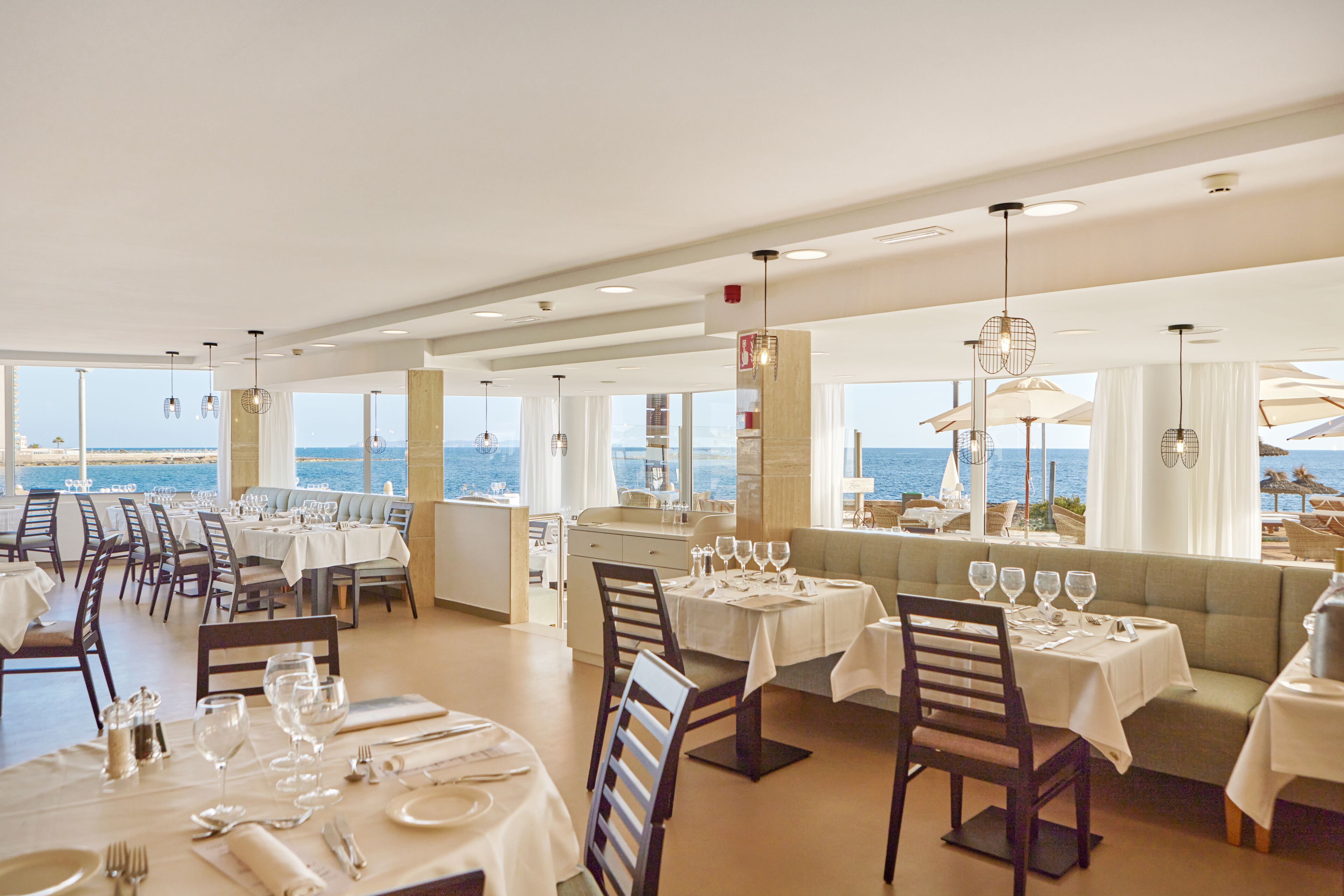 A restaurant with multiple tables and chairs, large windows with ocean views, and hanging pendant lights.