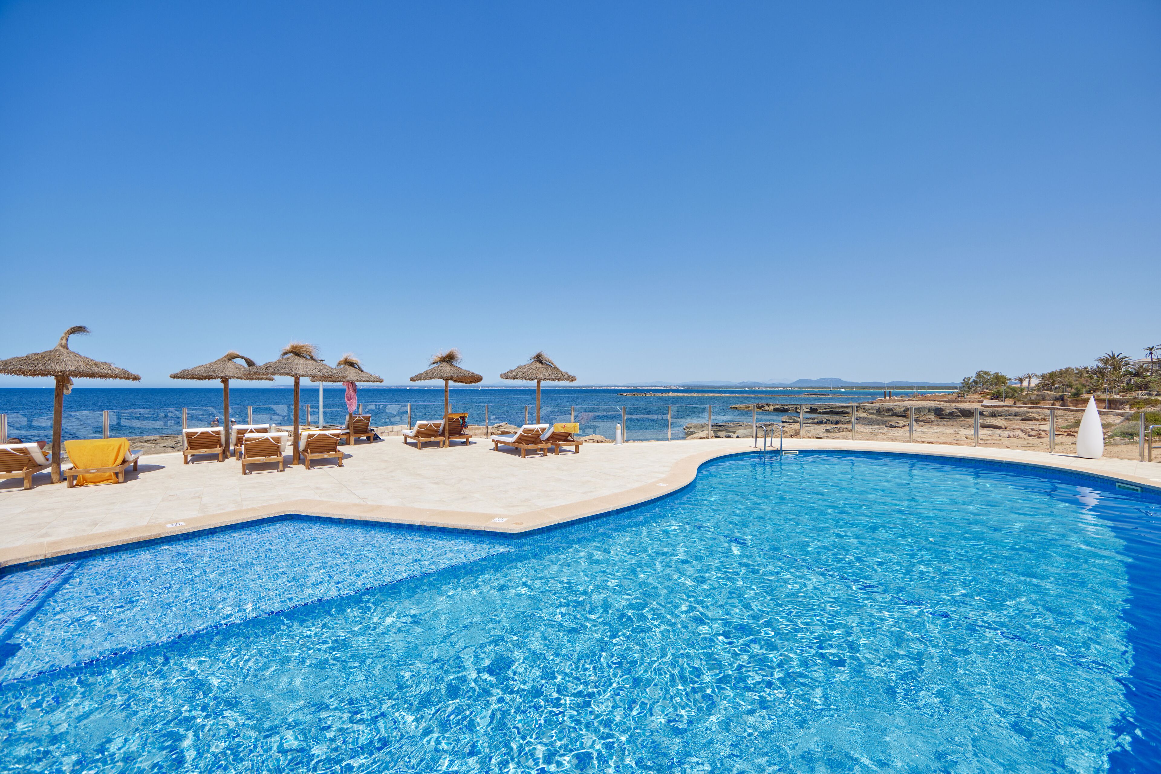 Une piscine bordée de chaises longues et d’ombrelles, avec vue sur une plage et l’océan.