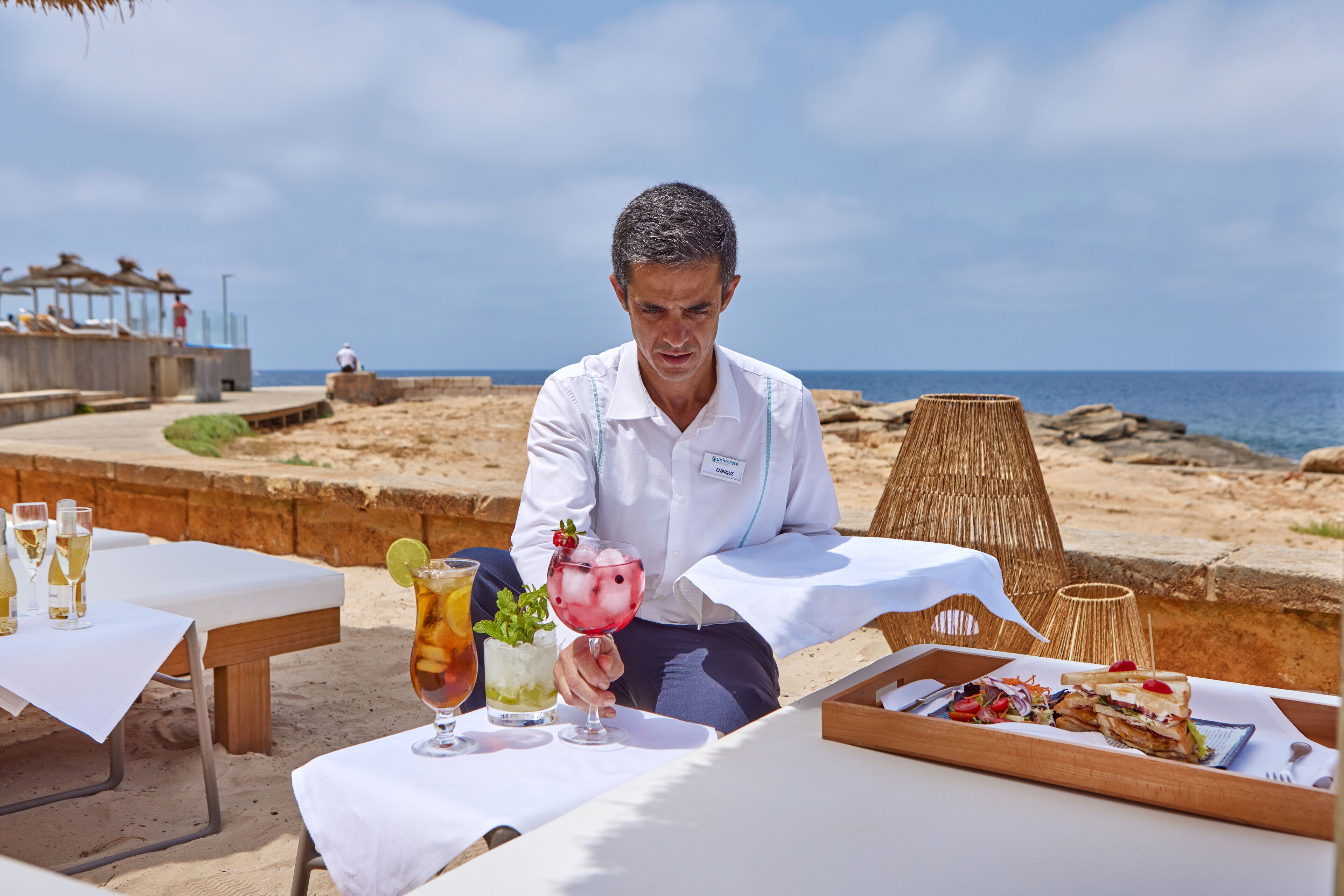 A beachside dining area with tables set for a meal, a drink menu, and a view of the ocean.