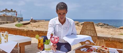 A beachside dining area with tables set for a meal, a drink menu, and a view of the ocean.