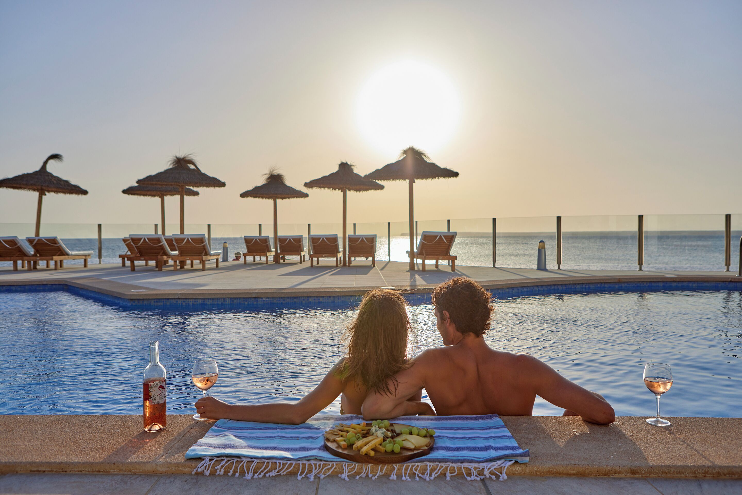 A couple relaxing by a poolside with a sunset view, lounge chairs, and umbrellas.