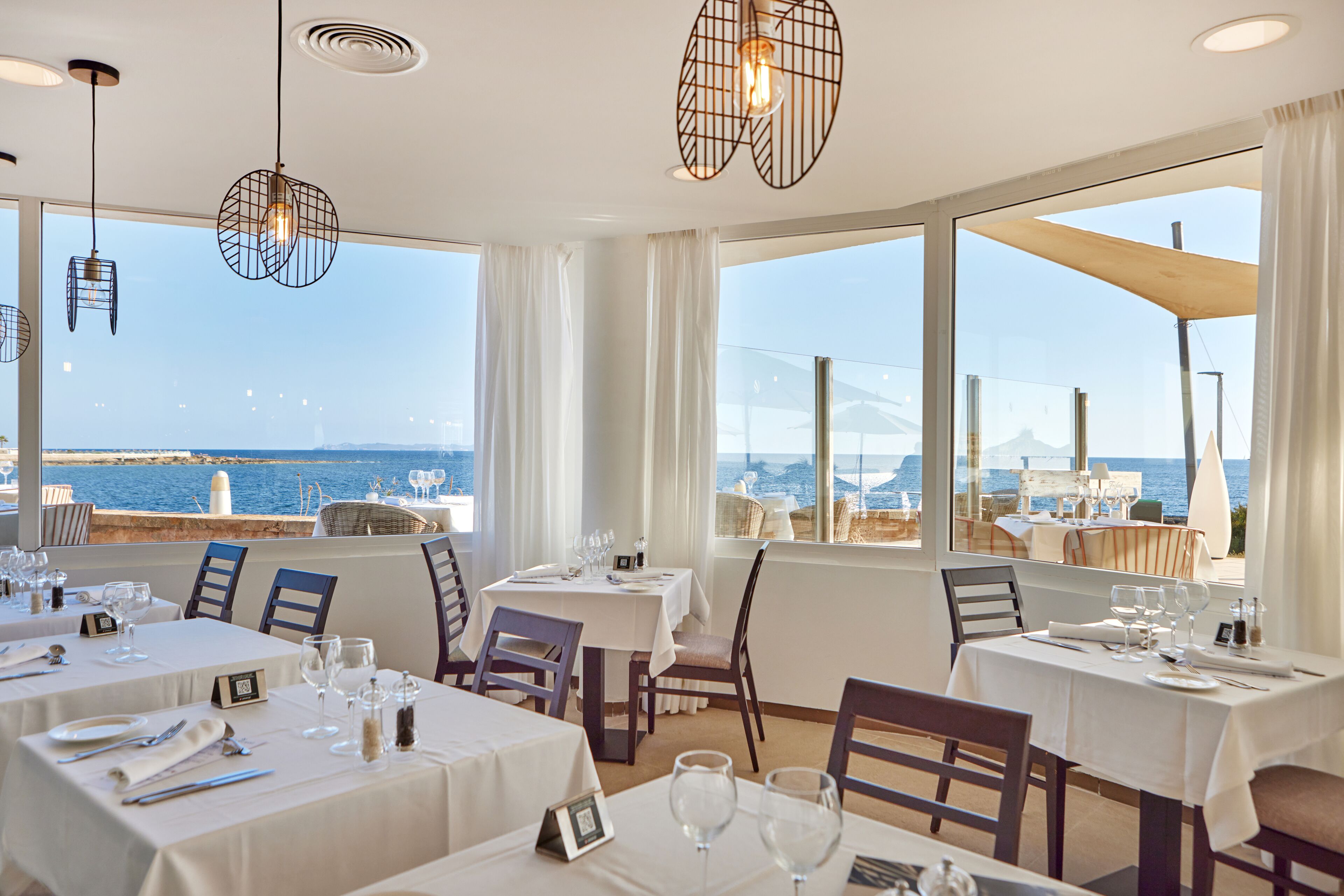 Un restaurant avec des tables dressées pour le service, de grandes fenêtres offrant une vue sur la mer et des lampes suspendues décoratives.