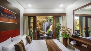 A hotel room with a bed, a small table with chairs, a potted plant, and a view of a waterfall through a glass door. - The Swaha Ubud Hotel (Batubulan)