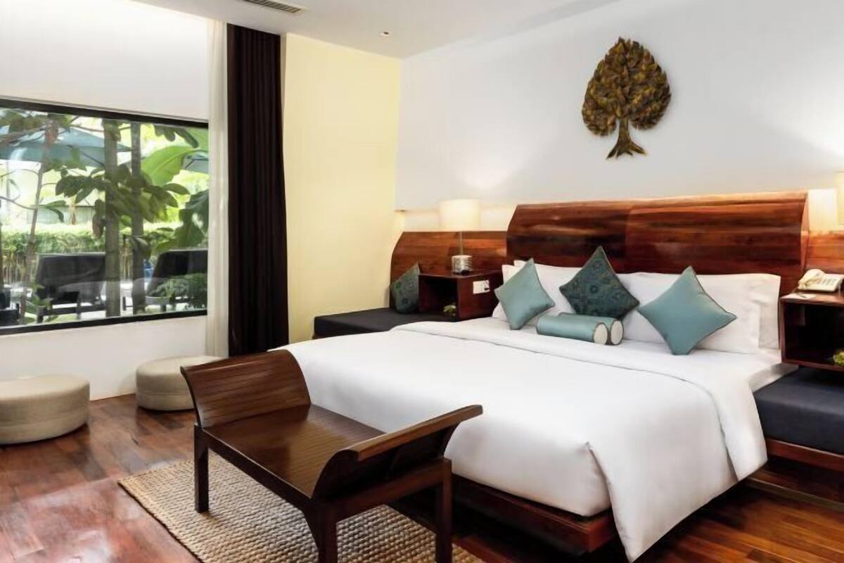 Une chambre d’hôtel avec un grand lit, un bureau en bois, une table de chevet et une fenêtre donnant sur un paysage verdoyant.