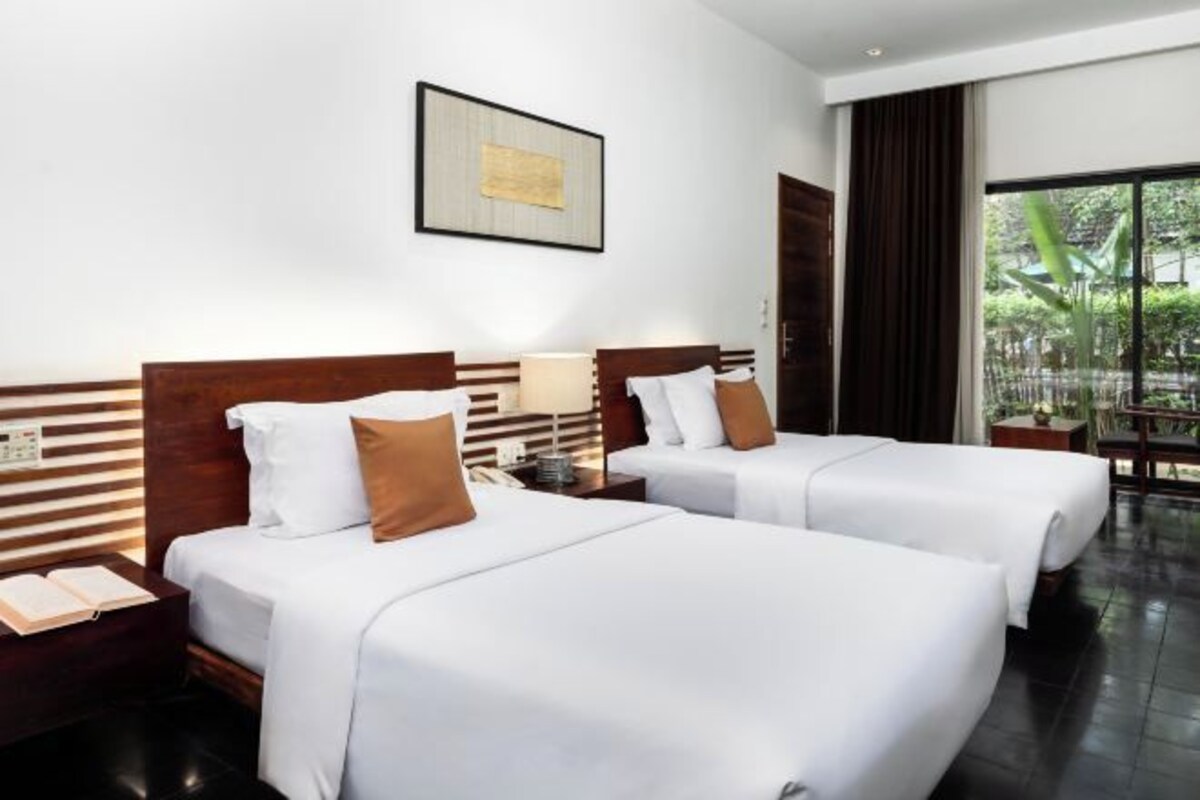 Een hotelkamer met twee bedden, een nachtkastje met een lamp, een lijst aan de muur en uitzicht op groen.