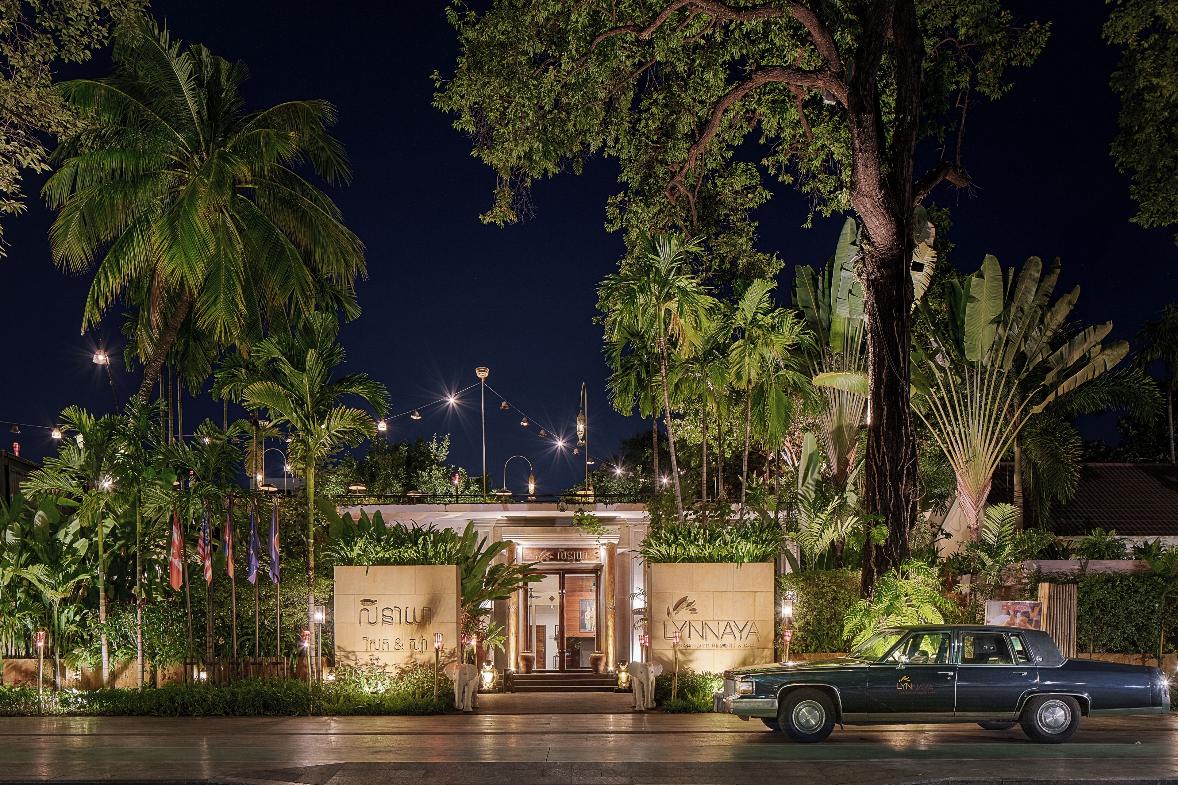 A hotel entrance with a car parked in front, surrounded by tropical vegetation and illuminated by string lights.
