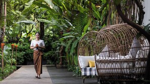 Une femme tenant une fleur dans un jardin luxuriant, avec une chaise en osier et un coussin à carreaux.