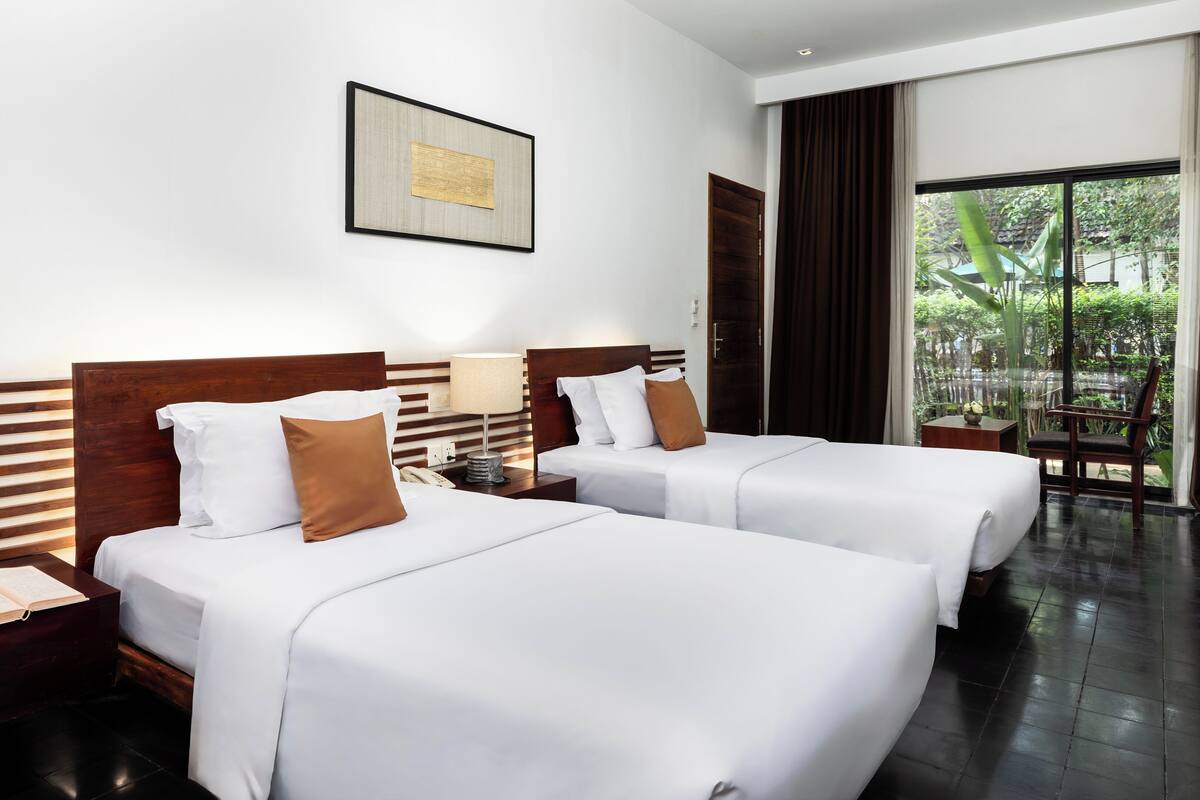 Une chambre d’hôtel avec deux lits, une table de chevet avec une lampe, un thermostat fixé au mur et une fenêtre donnant sur un paysage verdoyant.