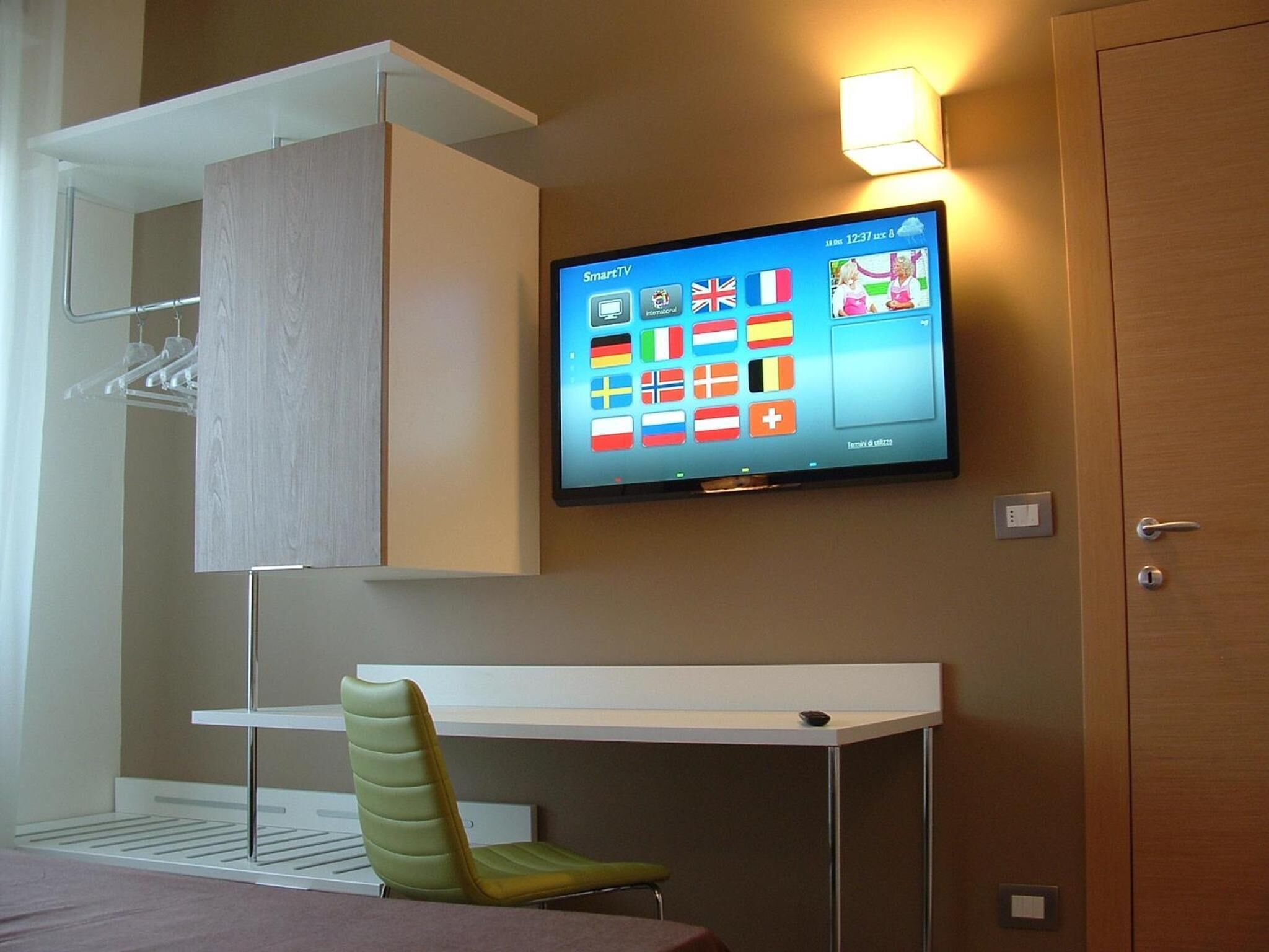 A hotel room with a flat-screen TV mounted on the wall, a desk with a chair, and a wardrobe with a hanging rack.