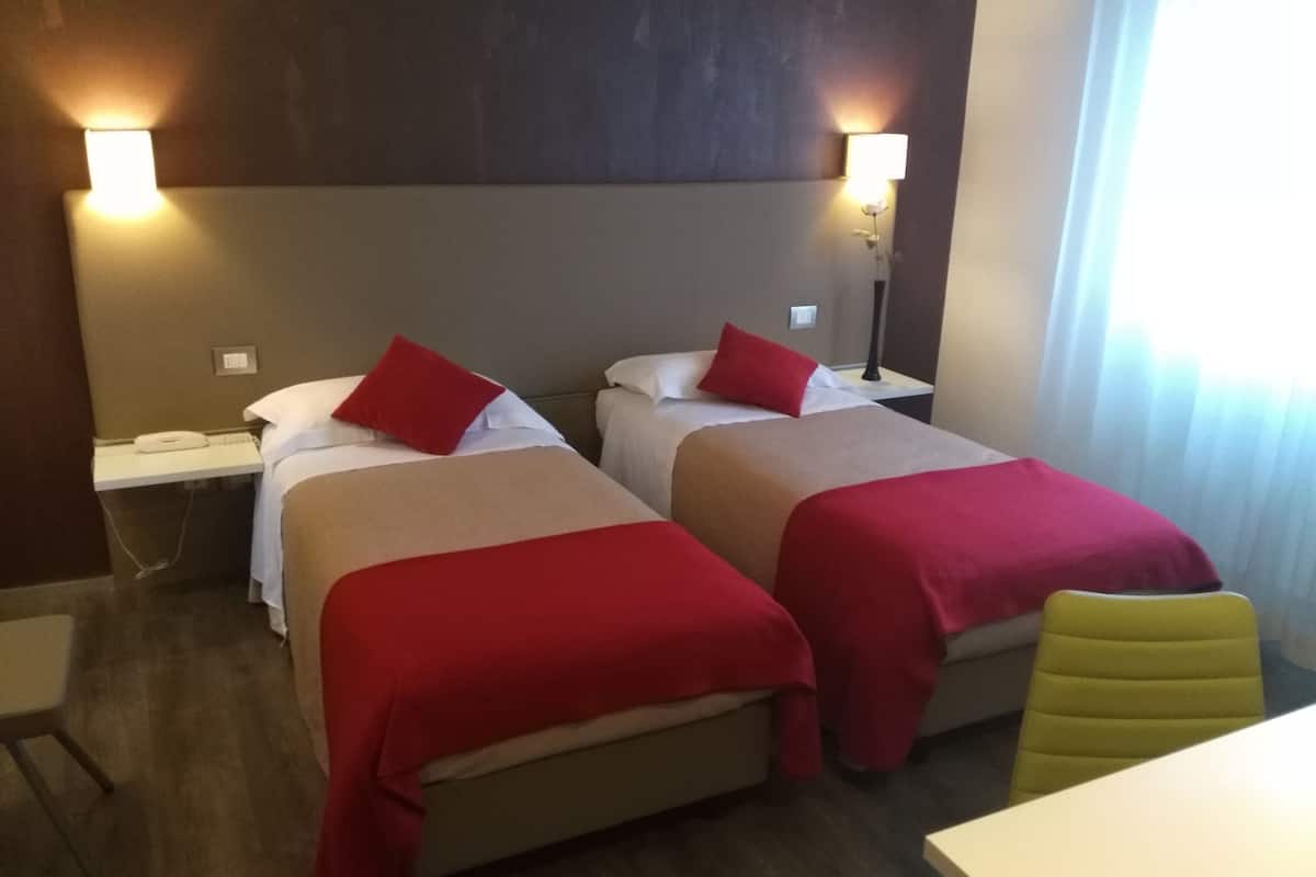 Een hotelkamer met twee bedden, elk voorzien van rode beddengoed en kussens, een nachtkastje met een lamp en een stoel.