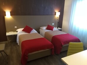Habitación de hotel con dos camas, cada una con ropa de cama y almohadas rojas, una mesita de noche con lámpara y una silla.