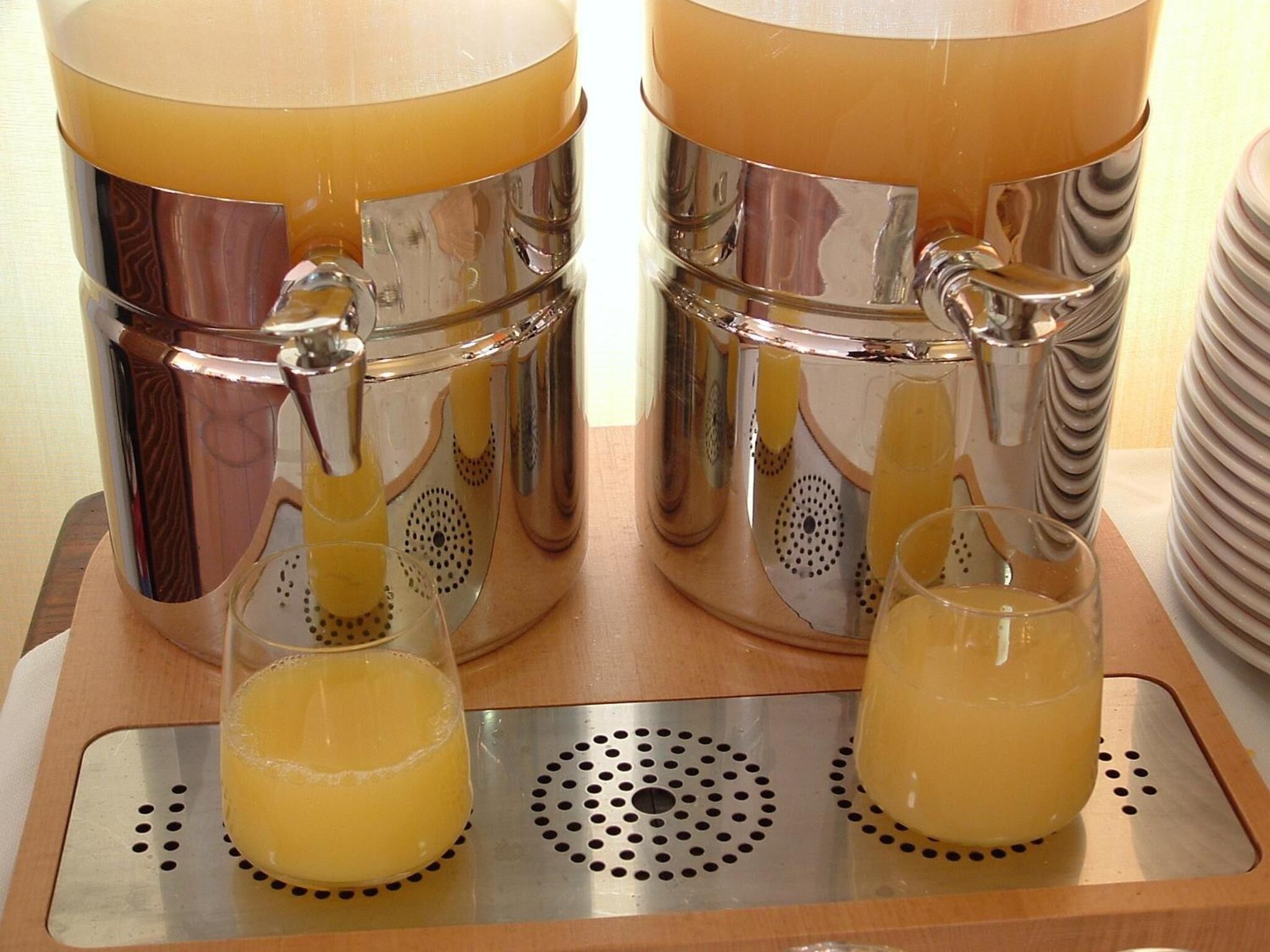 Two stainless steel beverage dispensers with spouts, two glasses filled with a yellow beverage, and a stack of plates.