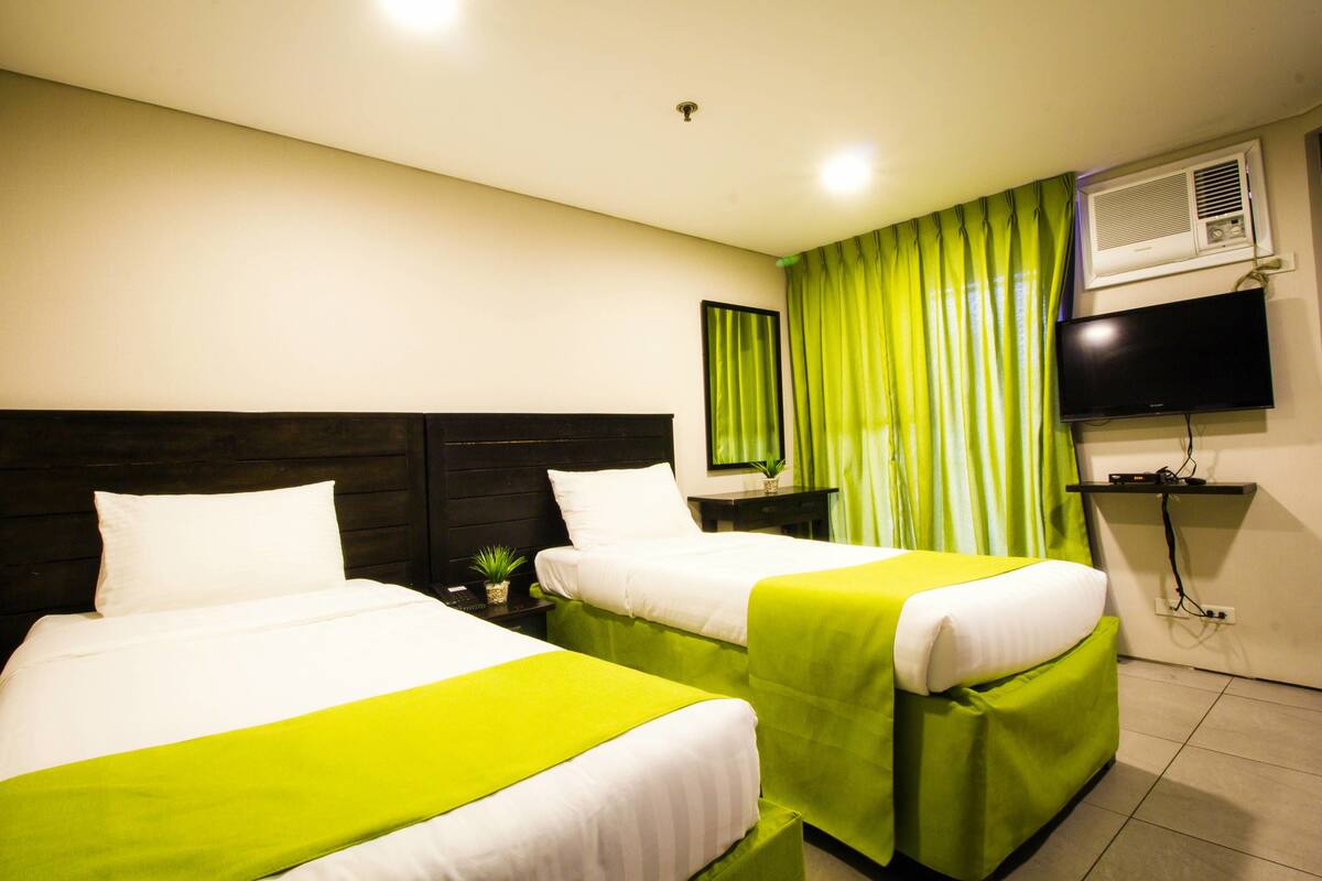A hotel room with two beds, a flat-screen TV, an air conditioning unit, and a green curtain.