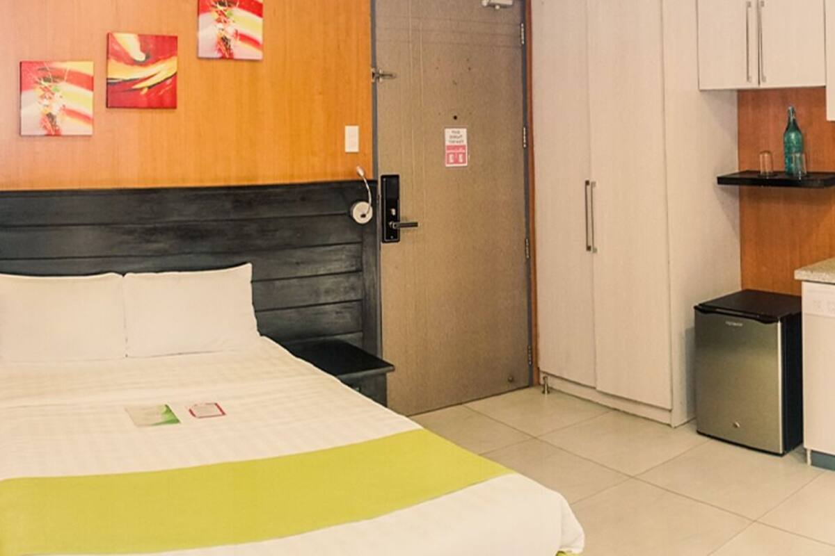 A hotel room with a bed, a small desk, a refrigerator, and a kitchenette.