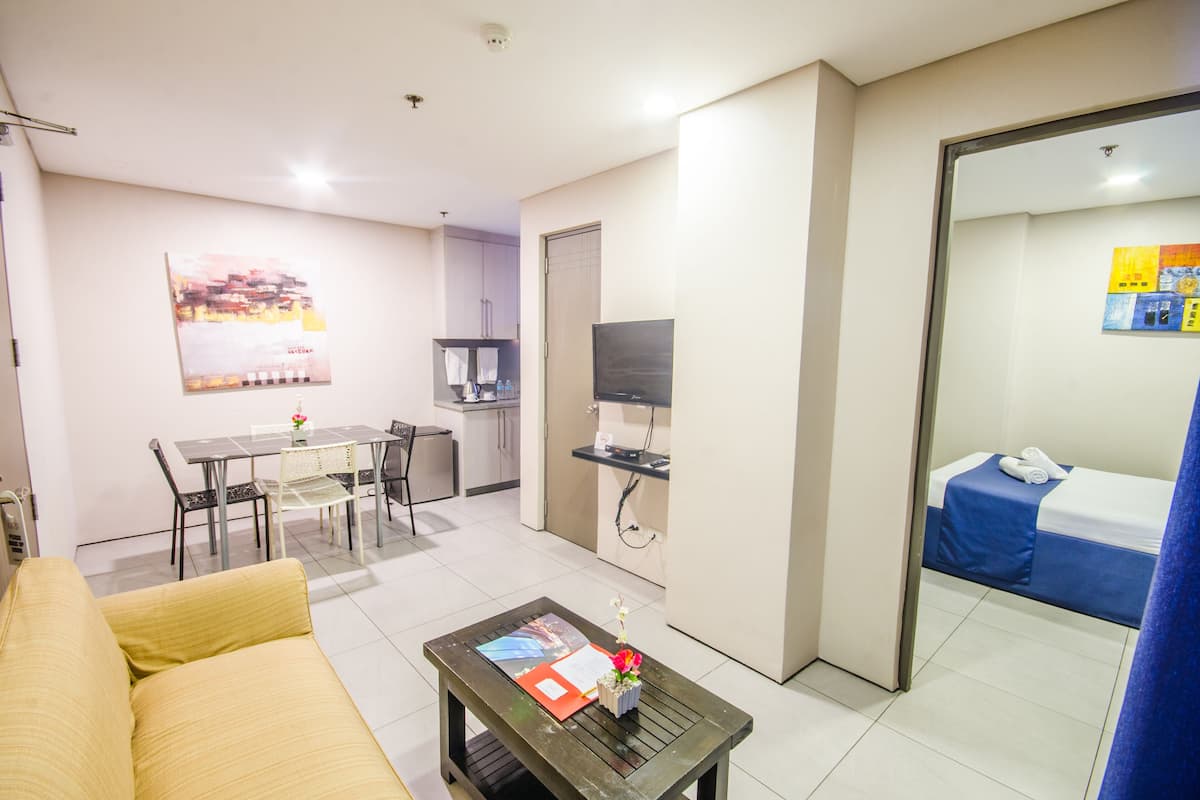 A compact living space with a sofa, a dining table with chairs, a small kitchen area, a TV, and a bed with a blue bedspread.