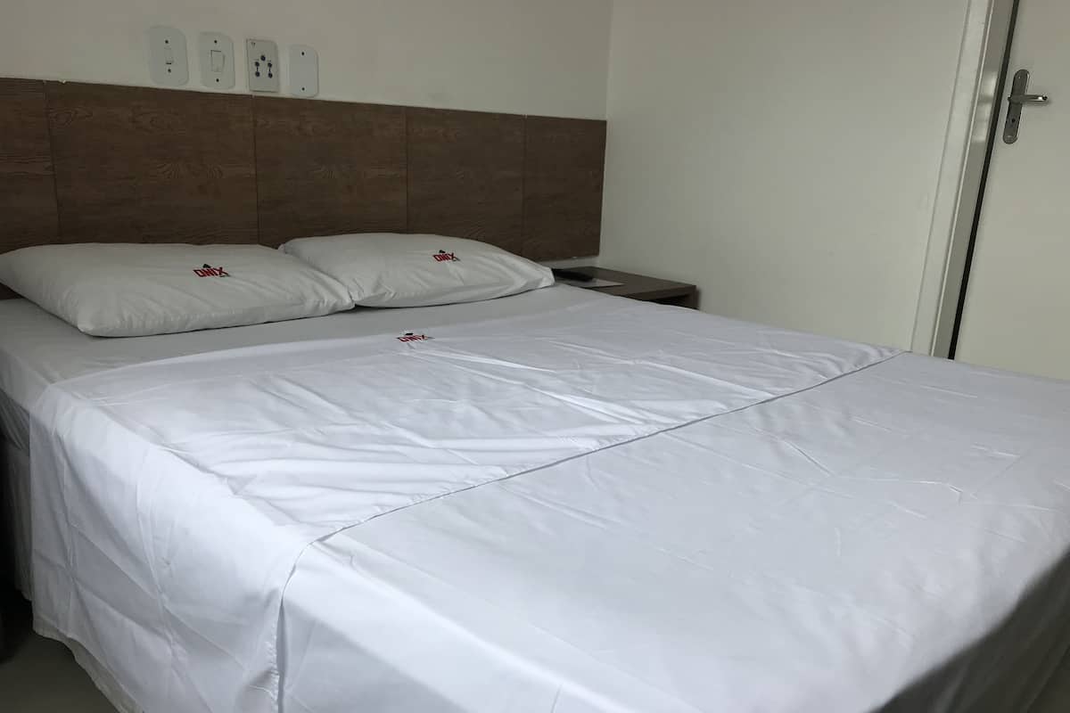 Ein ordentlich bezogenes Bett mit weißer Bettwäsche und zwei Kissen, ein hölzernes Kopfende, ein Nachttisch und eine Tür mit Griff.