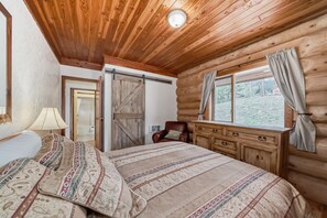 A log cabin bedroom with a bed, a chair, a dresser, and a window with curtains. - Terracana Ranch Resort (Valemount)