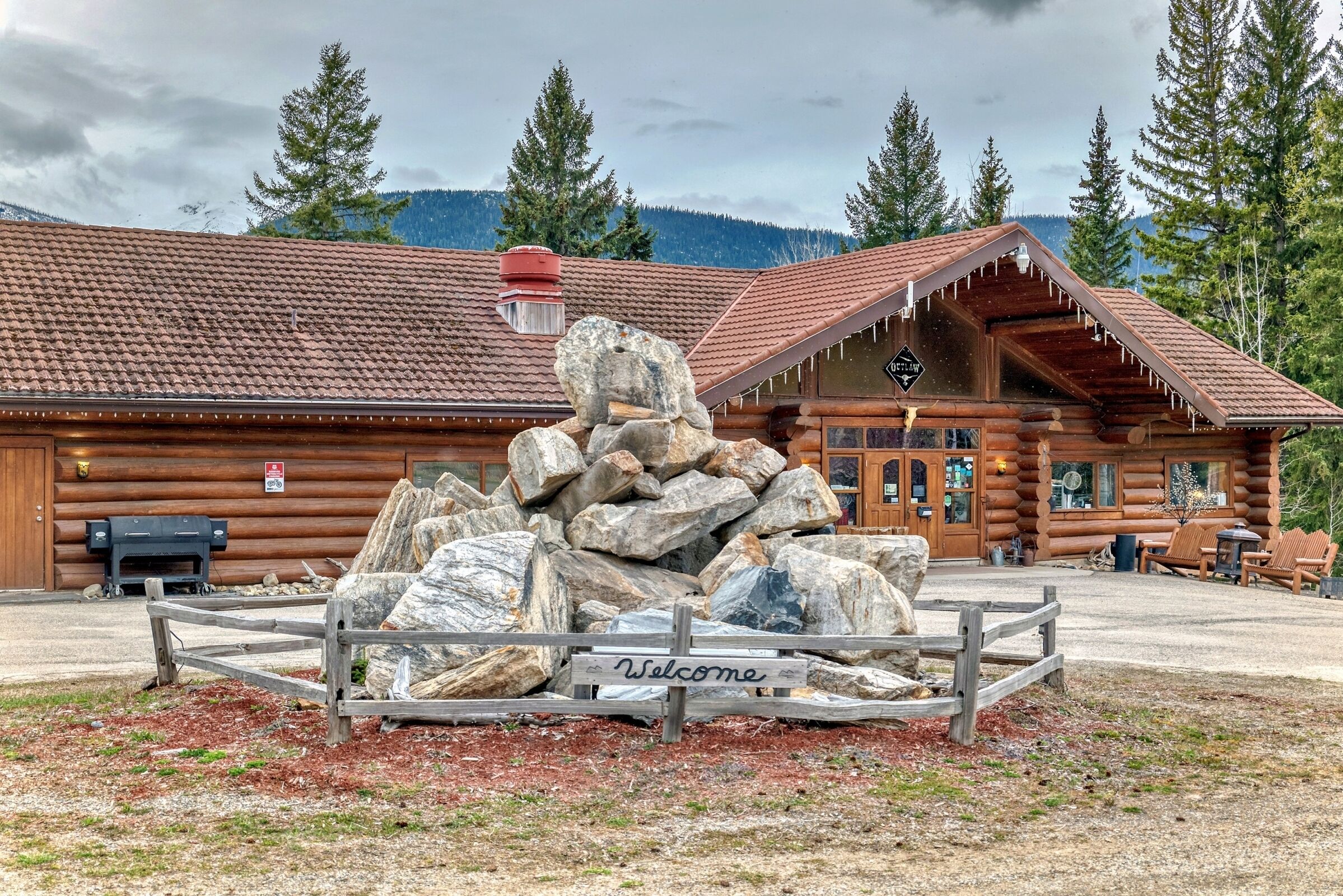 A log cabin with a 'Welcome' sign, a pile of rocks, and a mountain backdrop.