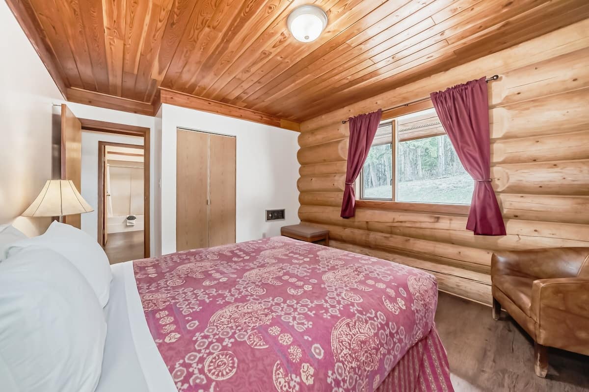 Un dormitori de cabana de fusta amb un llit, una tauleta de nit, una banca de fusta i una finestra amb cortines.