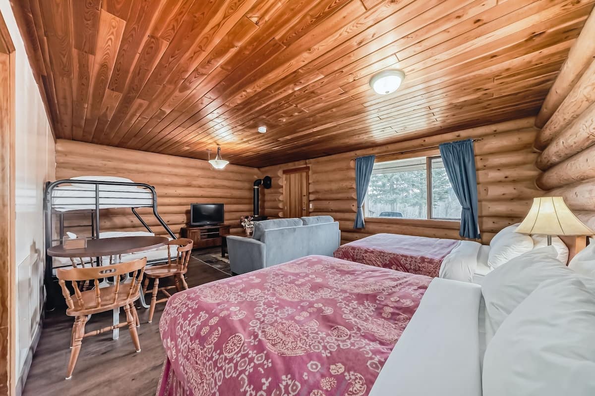 Habitació de cabana de fusta amb dos llits, una taula de dinar amb cadires, un televisor i una finestra amb cortines.