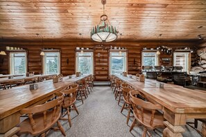 A log cabin interior with long wooden tables and chairs, a chandelier, and windows with views of trees and mountains.
