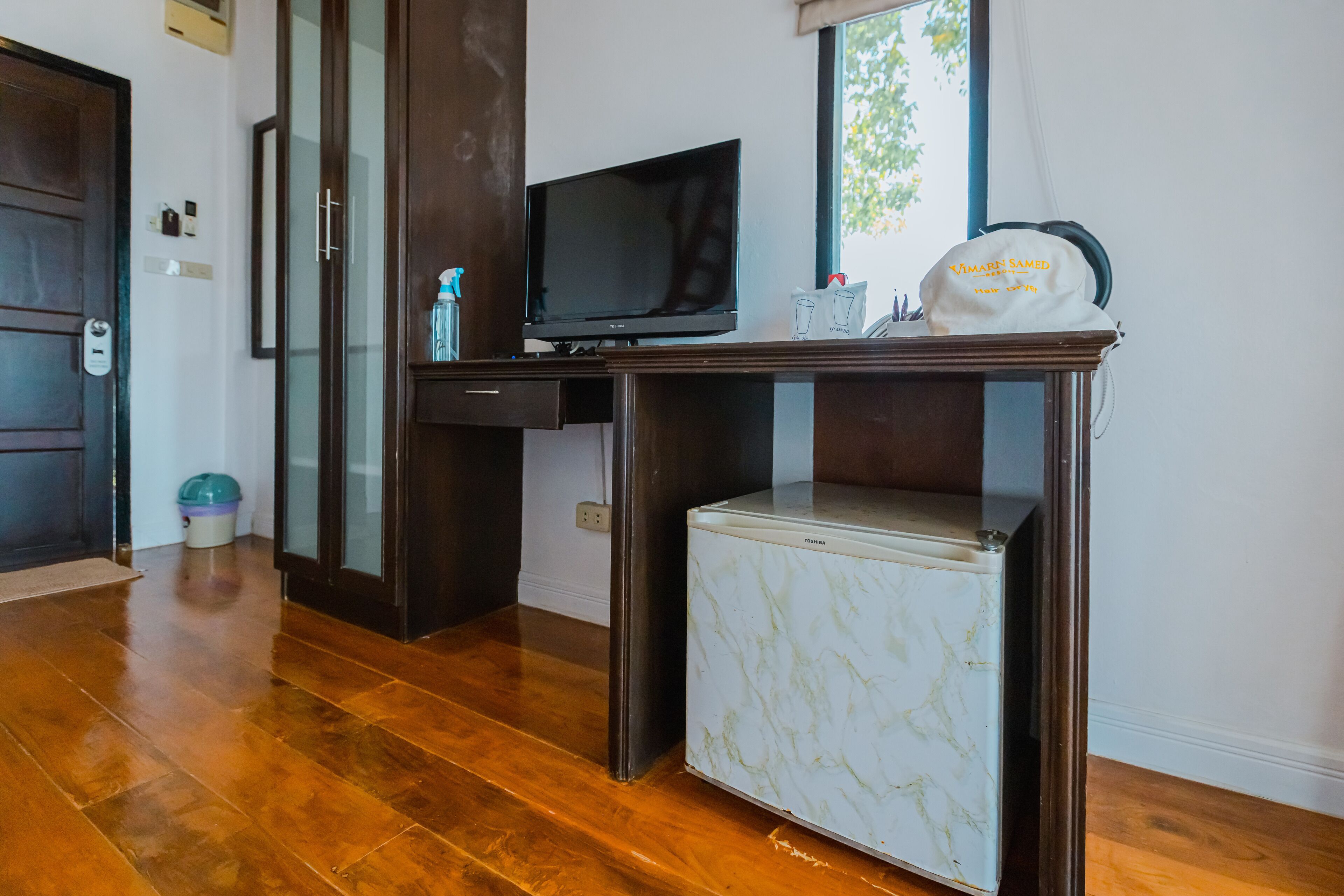 Habitación compacta con suelo de madera, un refrigerador blanco pequeño, un televisor de pantalla plana sobre un soporte de madera y una ventana con vista a zonas verdes.