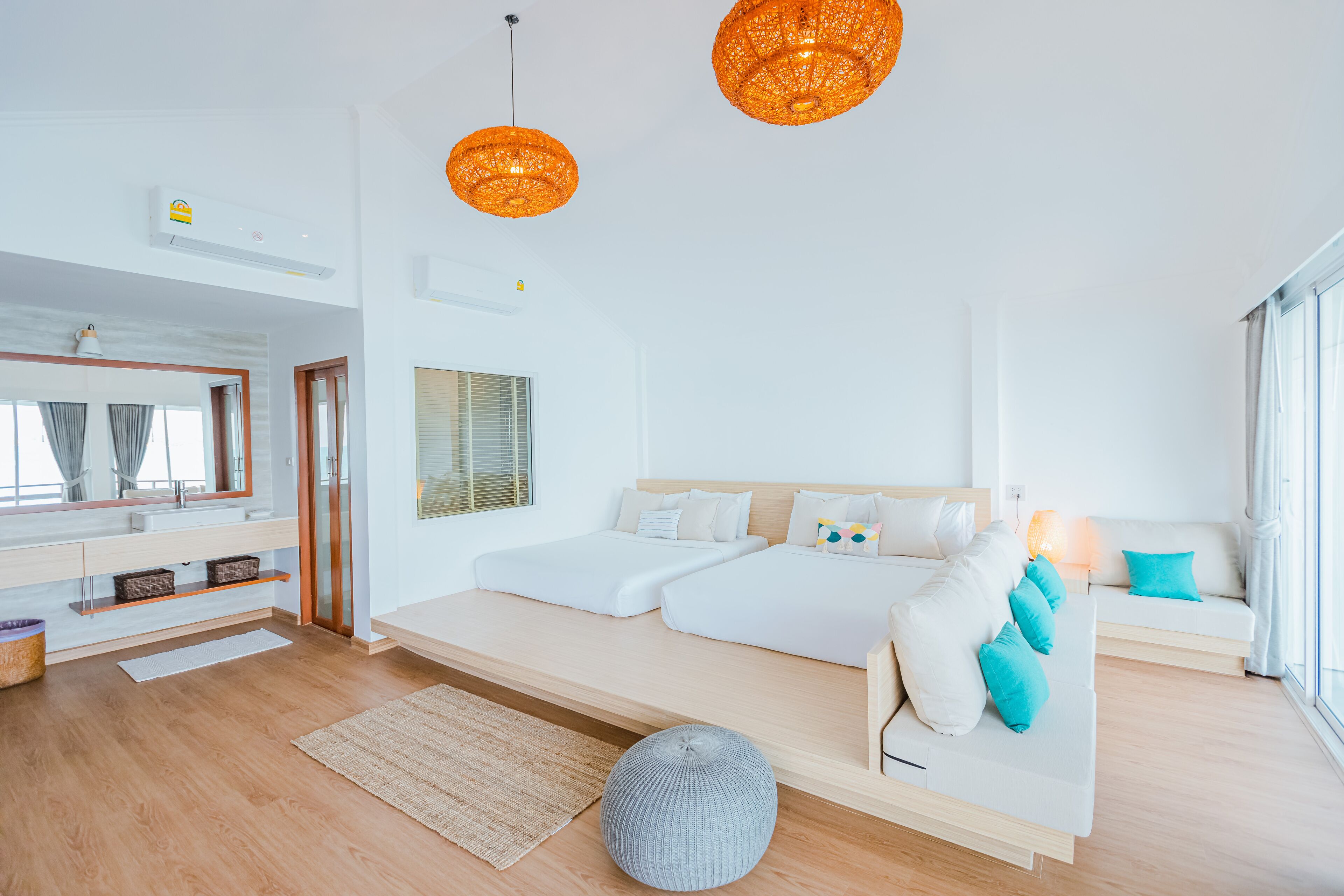 A modern bedroom with a large bed, a sofa, and a small ottoman. There is a window with curtains, a mirror, and a ceiling fan.