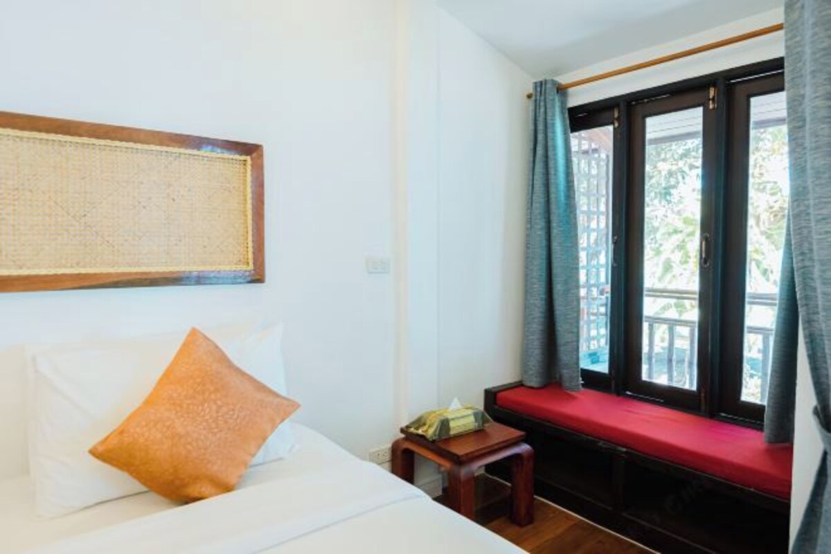 A hotel room with a bed, a small wooden table, a red bench by the window, and a view of greenery outside.