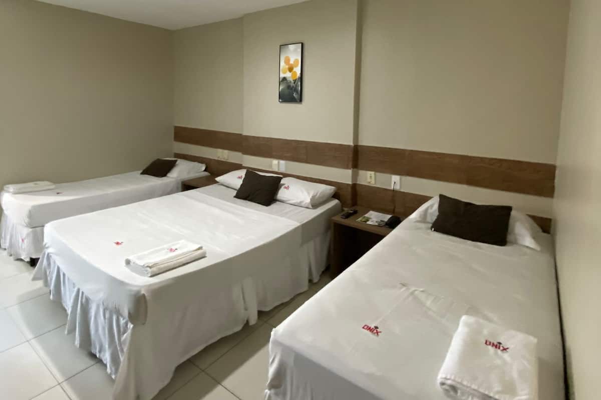 Habitación de hotel con tres camas individuales, cabecera de madera, mesita de noche pequeña con lámpara y un cuadro colgado en la pared.