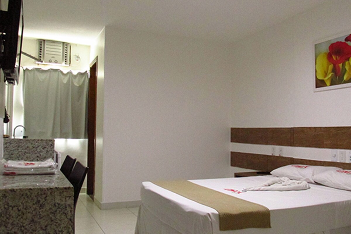Una habitació d'hotel amb un llit, una taula petita i una cadira. Hi ha un quadre a la paret i un aparell d'aire condicionat sobre el llit.