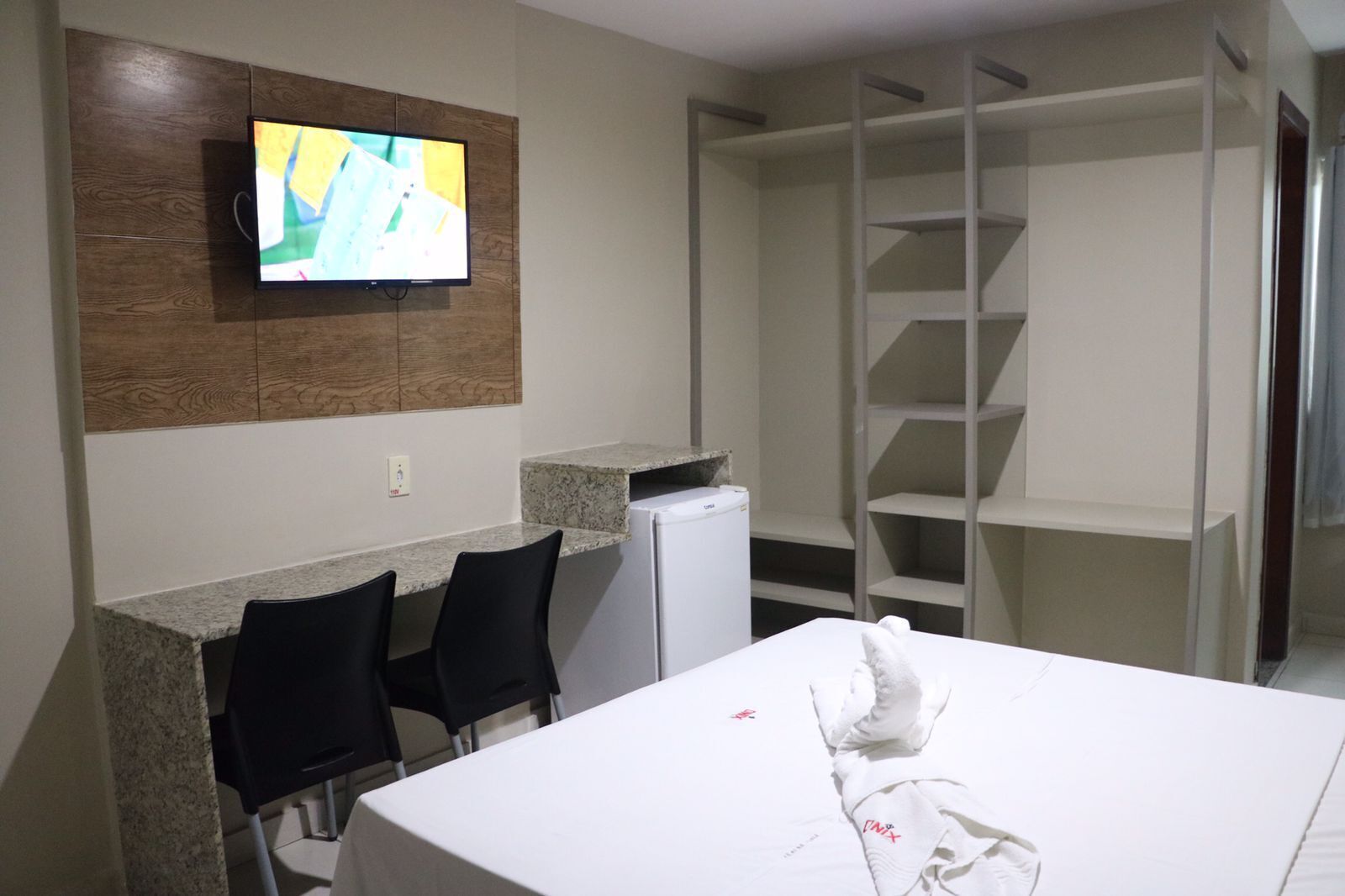 Habitació d'hotel amb un televisor fixat a un panell de paret de fusta, un llit blanc amb una tovallola blanca, un refrigerador petit, un taulell de granit i un moble d'emmagatzematge blanc.