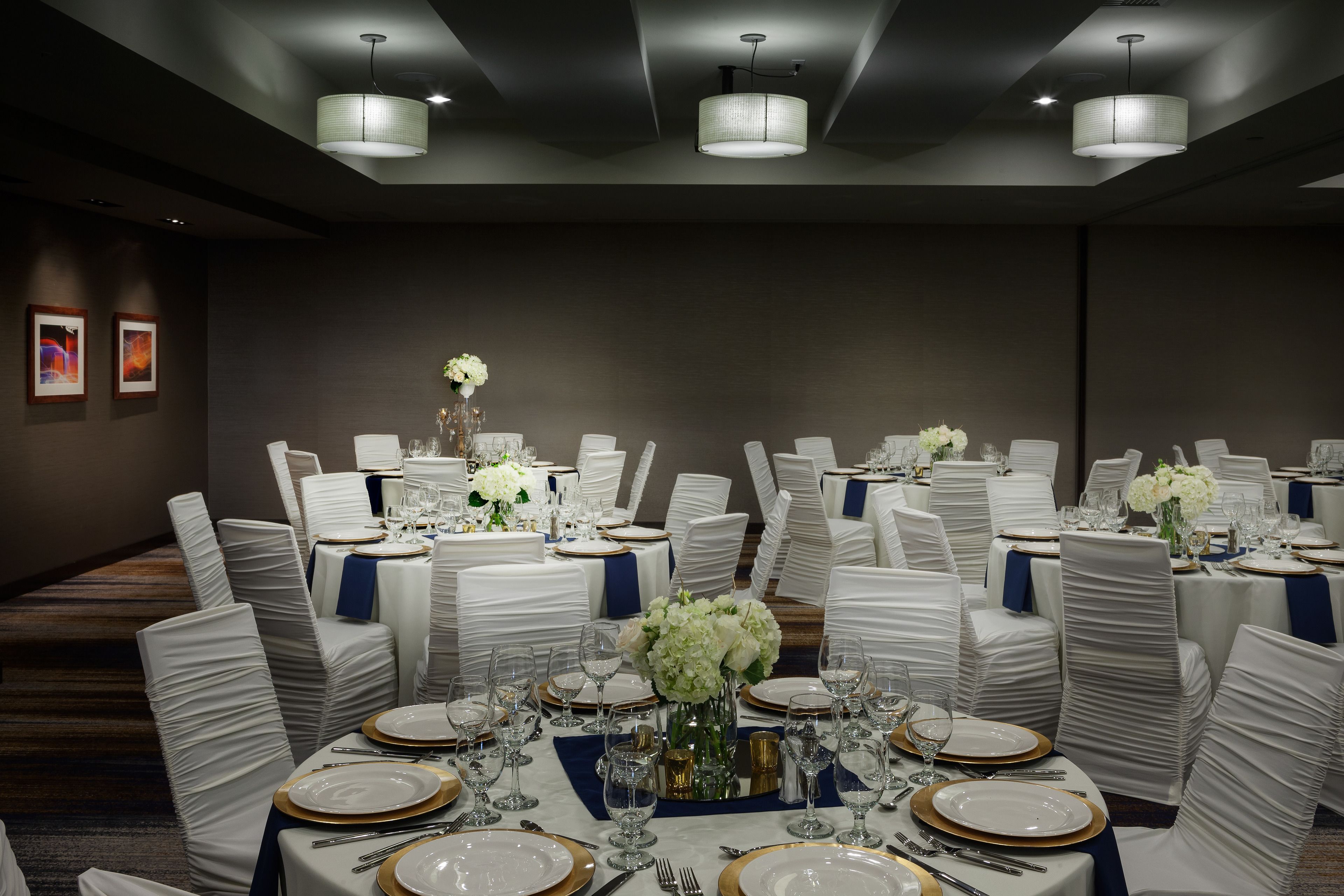 A banquet hall with round tables set for a formal event, featuring white tablecloths, gold-rimmed plates, and white chairs with blue sashes.