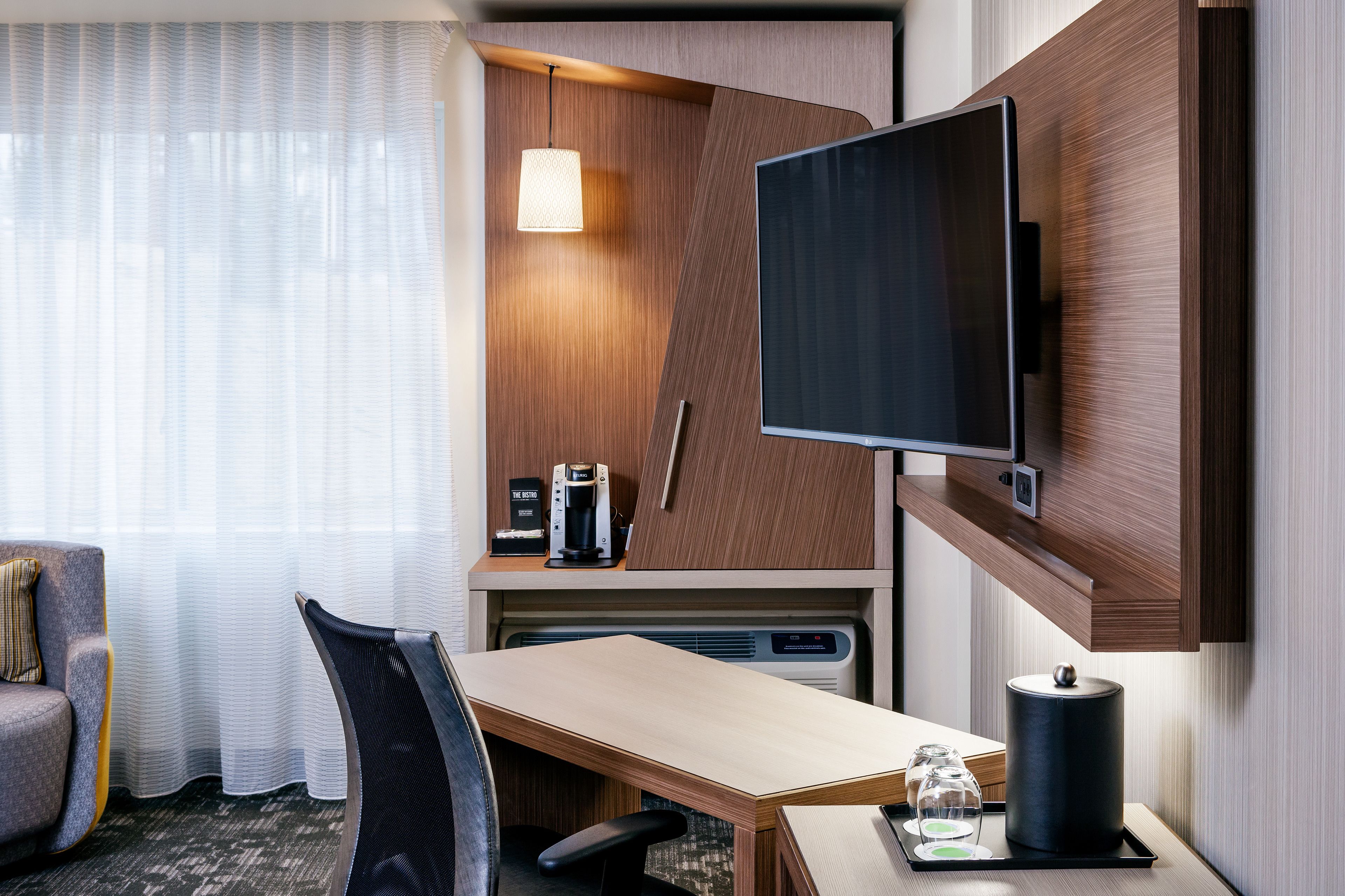 Une chambre d’hôtel avec un bureau en bois, un téléviseur fixé au mur, une chaise, une machine à café et une lampe.