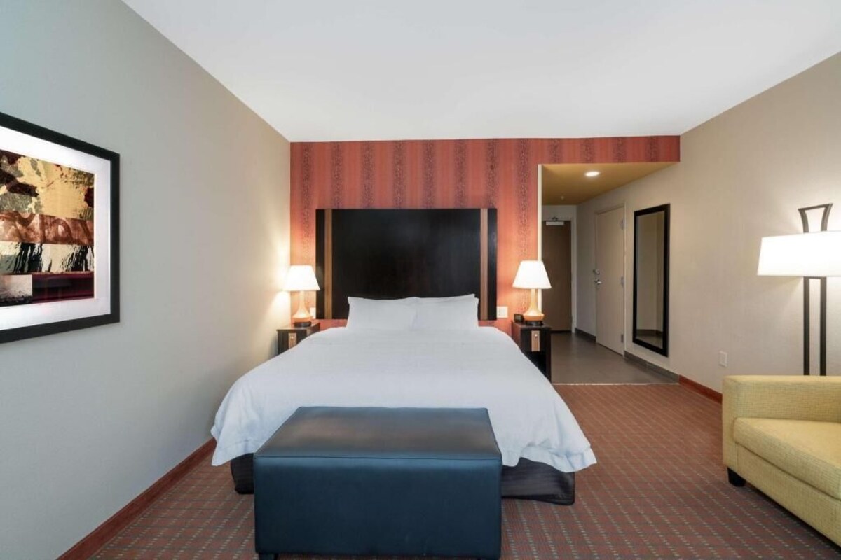 A hotel room with a bed, two bedside lamps, a large mirror, a framed picture on the wall, and a small bench.