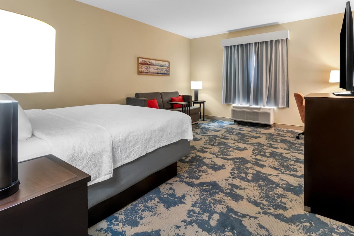 Habitación de hotel con cama, escritorio, silla, televisor, ventana con cortinas y un cuadro colgado en la pared.