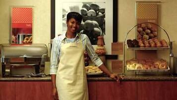 A person in a striped shirt and apron standing at a counter with a variety of baked goods and a coffee machine.