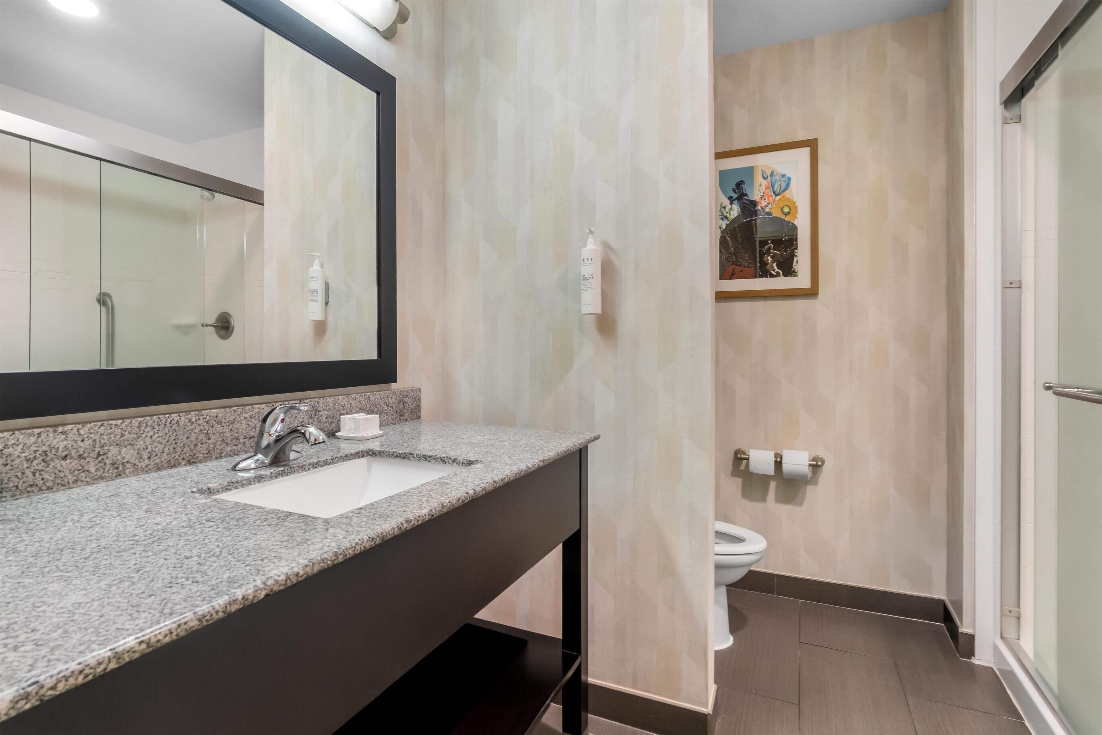 Une salle de bain moderne avec un lavabo surmonté d’un plan de travail en granit, un grand miroir, des toilettes et un espace douche.