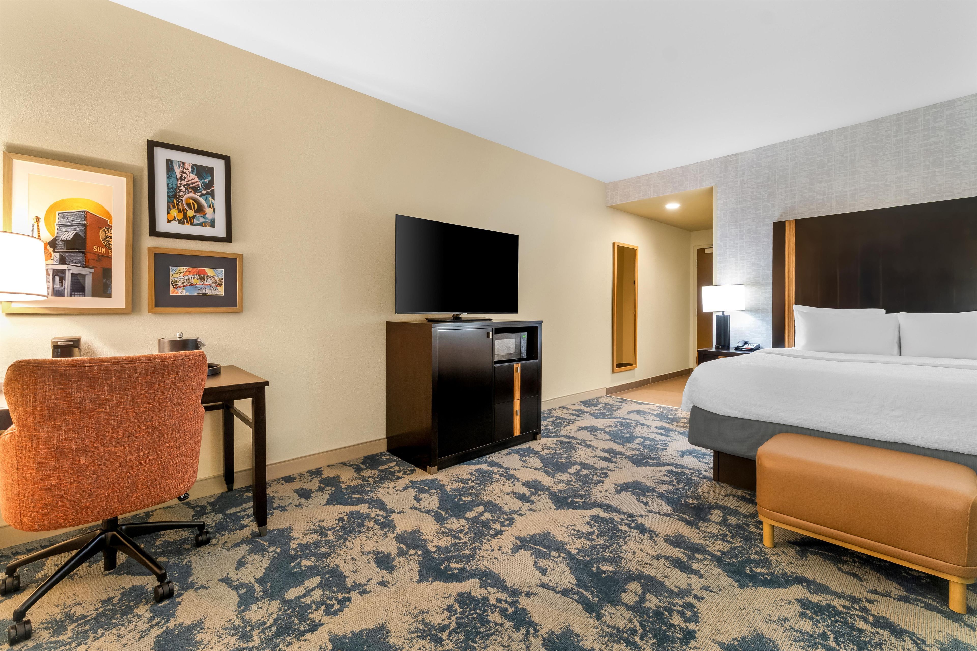 Habitación de hotel con cama, escritorio, silla, televisor y una obra de arte en la pared.