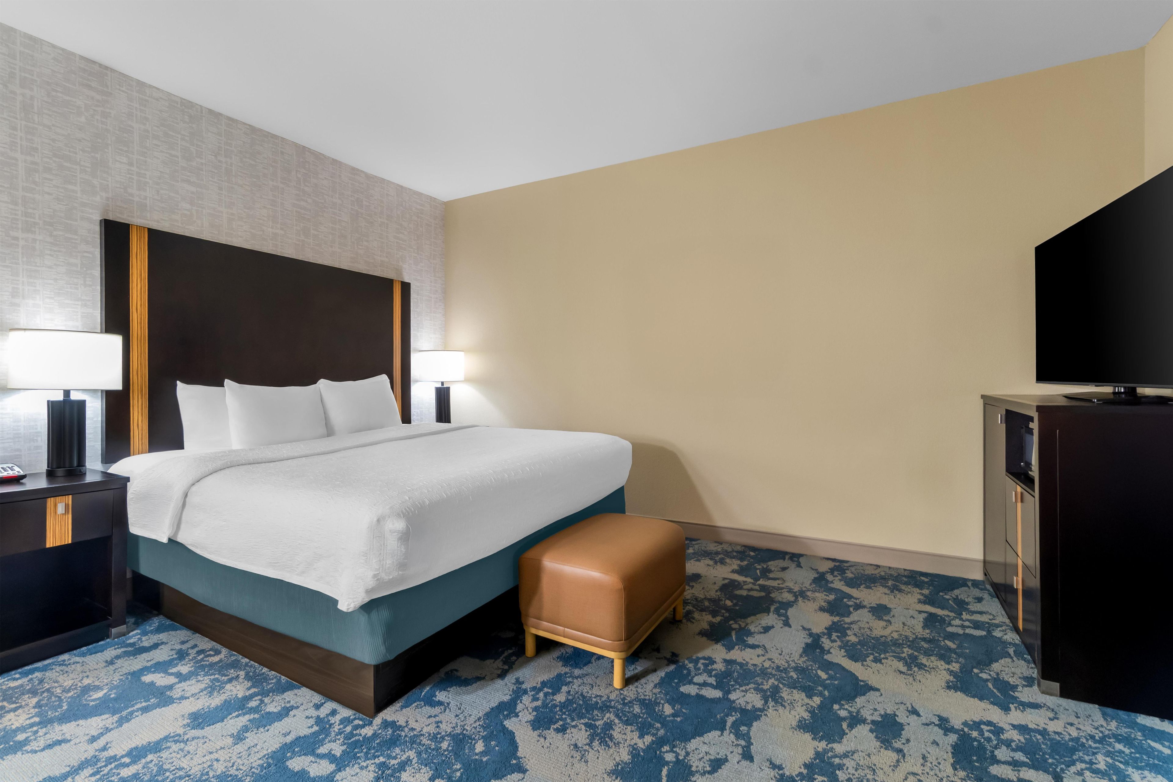 A hotel room with a bed, bedside lamps, a desk, a television, and a patterned carpet.