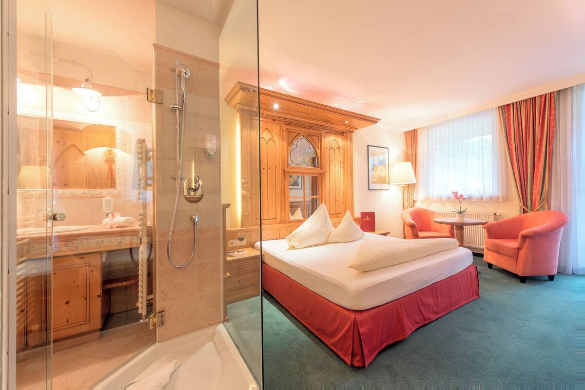Habitación de hotel con cabina de ducha de vidrio, cama con cubrecolcha rojo, aparador de madera, tocador con espejo y una mesita con lámpara.