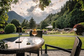 Een terras met een ronde tafel en stoelen, een glas witte wijn en een cocktail met rietje.
