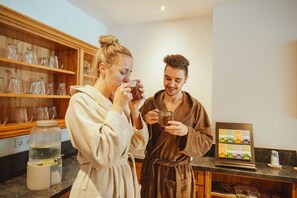 Deux personnes en peignoir se trouvent dans une cuisine équipée d’un meuble en bois, d’une machine à café et d’un tableau encadré posé sur le comptoir.